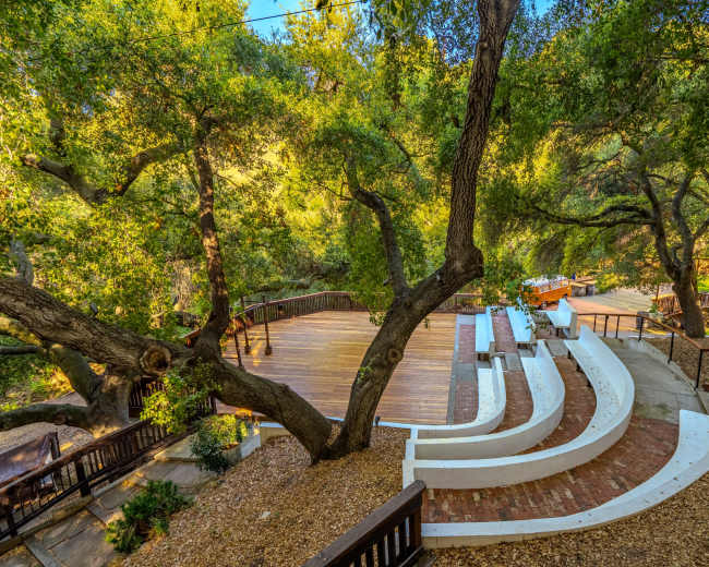 The image shows an outdoor amphitheater surrounded by trees, with tiered seating and a wooden stage area in a natural setting.