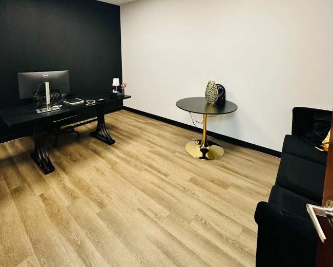 The image shows a small office space featuring a black wall, a desk with a computer, a round table, and a couch on a wood floor.