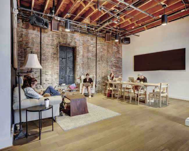 The image shows a spacious, well-lit room with exposed brick walls, featuring a seating area with a sofa and a coffee table, and a long table where several people are working on laptops.