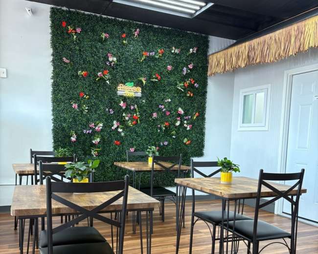 The image depicts a cozy cafe interior featuring several metal-framed tables with black chairs, surrounded by a green wall adorned with flowers.