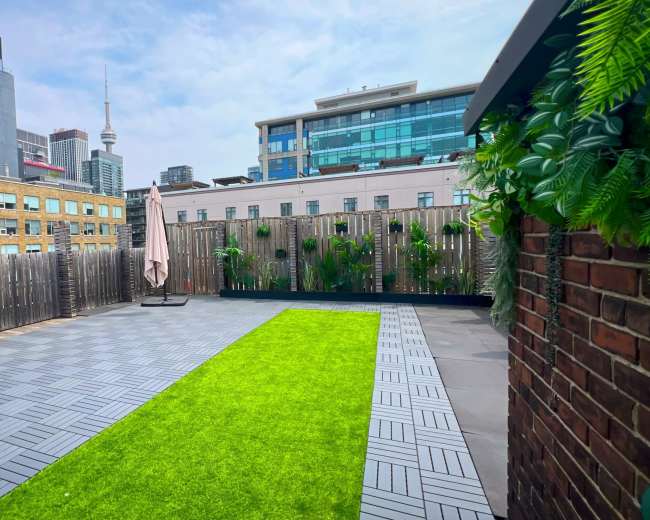 The image shows a rooftop terrace with artificial grass, surrounded by wooden fences and city buildings in the background, including a visible tower.