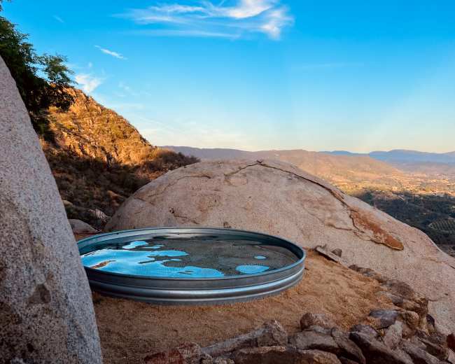 A circular metal dish sits on a rocky surface, reflecting a clear sky and surrounding landscape.