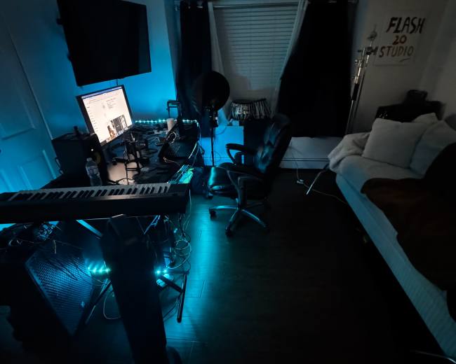 The image shows a dimly lit room featuring a computer setup, a keyboard, and a bed, with blue lighting casting a cool tone throughout the space.