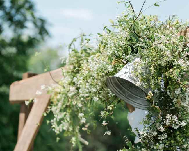 A metal bucket overflows with greenery and small white flowers, hanging from a wooden structure outdoors.