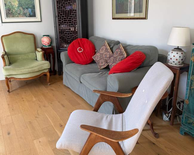 The image depicts a living room with a gray sofa, accented by red pillows, a wooden chair, and various framed artworks on the walls.