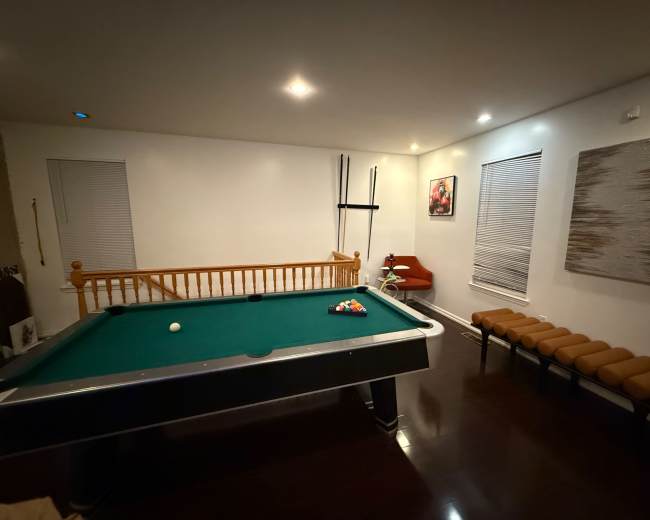The image shows a billiards room featuring a green pool table, a wooden railing, and a seating area with a red chair and a long bench.
