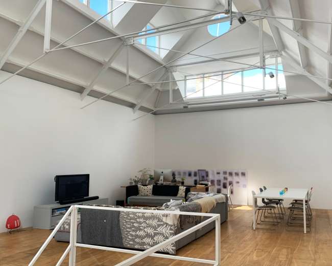 The image shows a spacious, modern living room with a high, vaulted ceiling, a wooden floor, a sectional couch, a television, and a dining table with chairs.