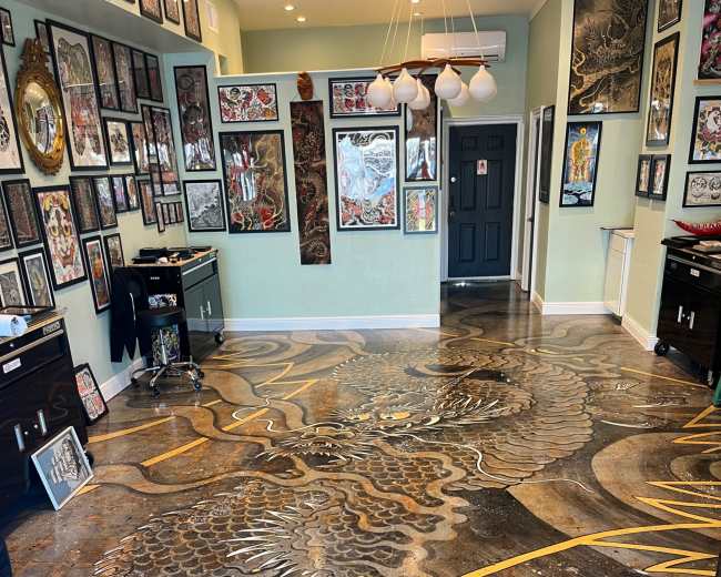 The image shows a tattoo studio with an intricately designed floor featuring a dragon motif, surrounded by numerous framed artworks on the walls.