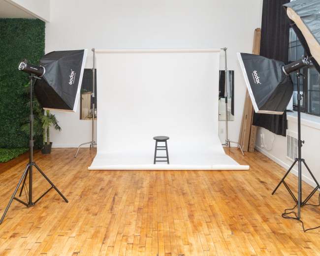 A photography studio features a white backdrop with a black stool in the center, surrounded by two lighting setups.