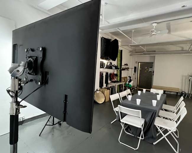 A large monitor is positioned beside a rectangular table covered with a gray tablecloth and surrounded by white chairs in a spacious, well-lit studio.