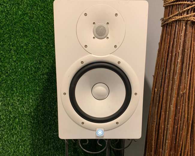 A white speaker stands on a decorative metal stand in front of a backdrop of artificial grass and a tall bundle of branches in a pot.