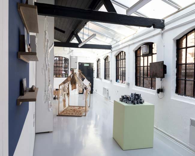 The image shows a spacious art gallery with large windows, white walls, and various installations, including a wooden structure and grouped objects on a pedestal.