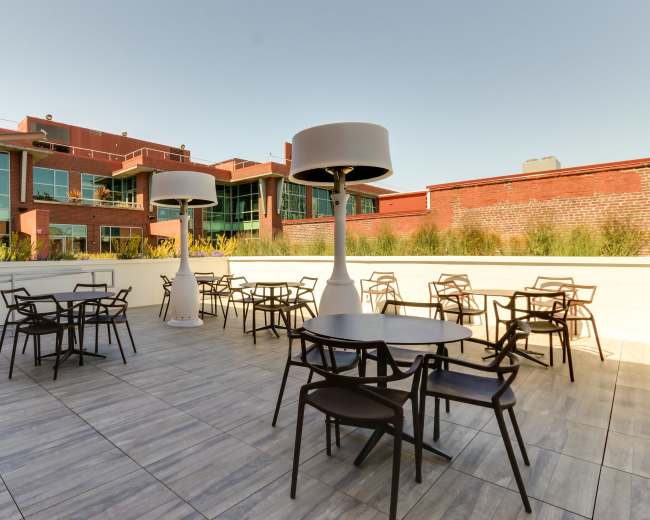 The image shows a spacious outdoor terrace featuring several round tables and oversized lamps, bordered by a building with large glass windows.