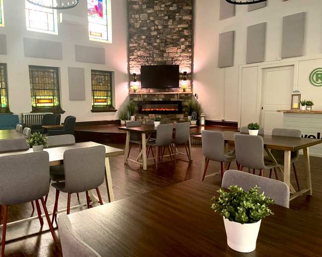 A spacious, well-lit room with multiple tables arranged around a modern fireplace and stained glass windows.