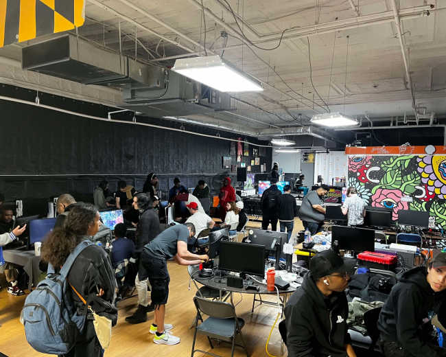 A crowded gaming center features multiple people engaged with computer setups and screens, while others socialize in a vibrant environment decorated with colorful murals.