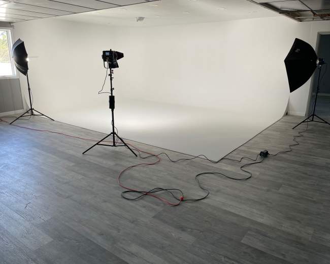 A photography studio with a seamless white backdrop, two light sources on tripods, and cables running across a wooden floor.