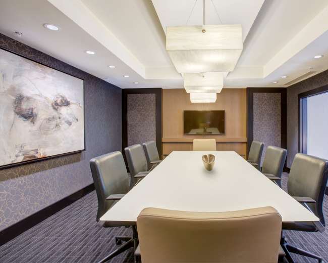 A modern conference room features a long white table surrounded by leather chairs, with overhead pendant lights and artwork on the walls.