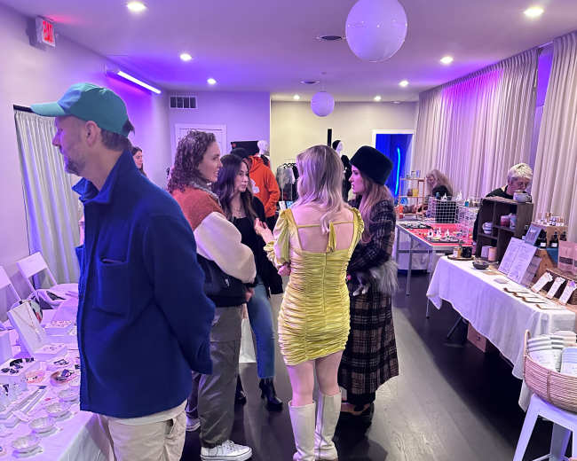 A group of people interacts at a brightly lit event space featuring tables with various goods on display.