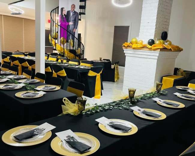 A dining area is set with black and gold table settings, featuring plates, utensils, and decorative elements, alongside a staircase adorned with balloons.