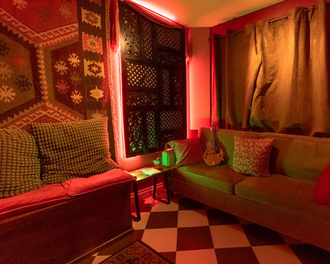 The room features a cozy seating area with a guitar resting on a couch, surrounded by patterned textiles and softly illuminated by red lighting.