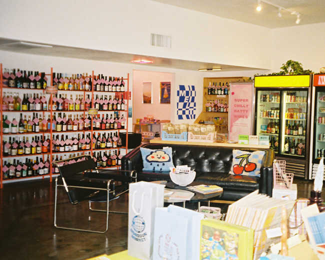 The image shows a brightly lit store interior featuring colorful shelves of beverages, a black seating area, and a display of books and bags on tables.