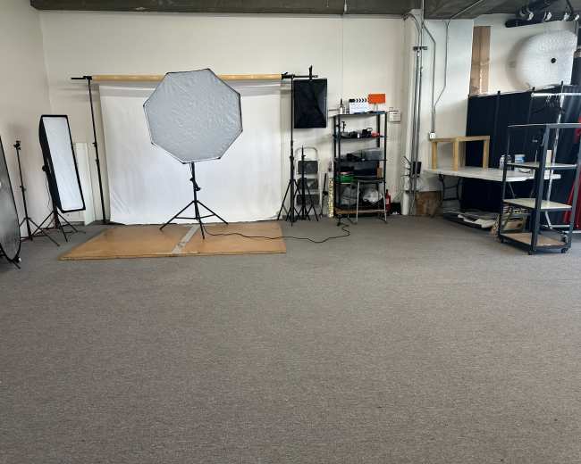 The image shows an empty photography studio with a white backdrop, an octagonal softbox light, and various equipment arranged on shelves.