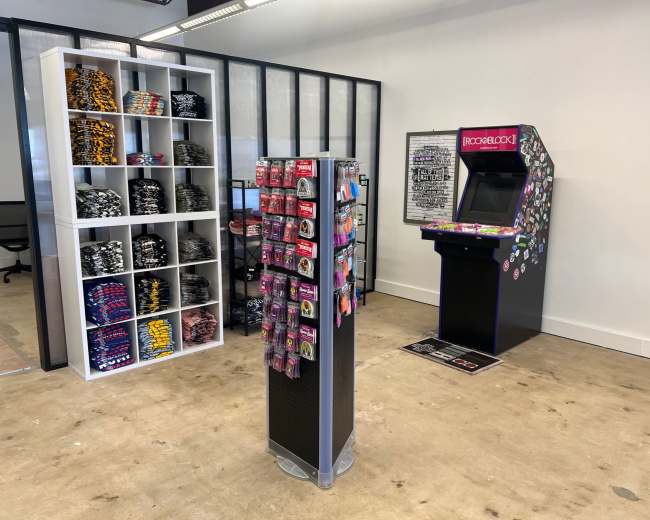 The image shows a retail space featuring a display of colorful merchandise on shelves, a stand with various items, and an arcade machine in the corner.