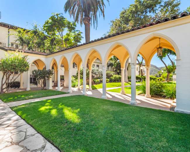 The image shows a colonnade with arches leading to a landscaped garden surrounded by greenery and palm trees.