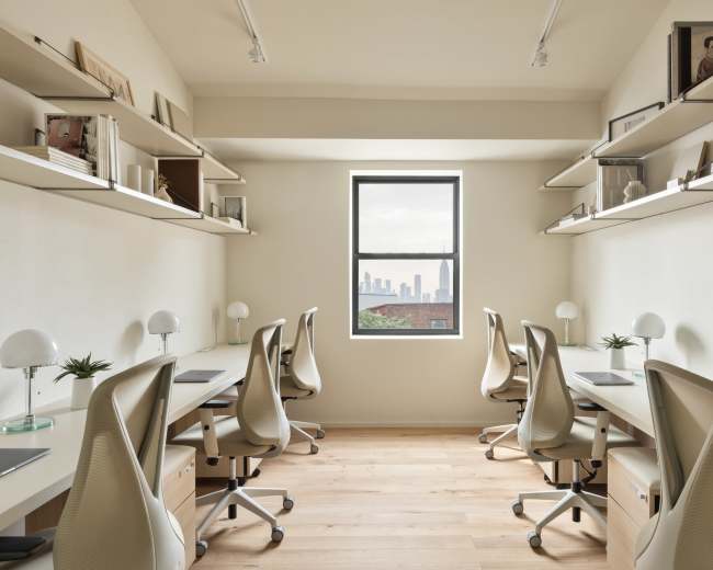 A modern office space features six workstations with chairs, shelves filled with books and decor, and a large window overlooking a city skyline.