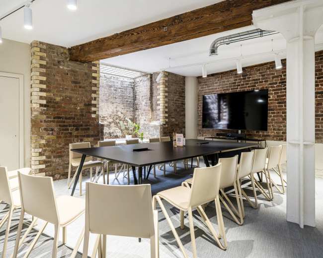 A conference room features a long black table surrounded by beige chairs, with exposed brick walls and a large screen on one side.