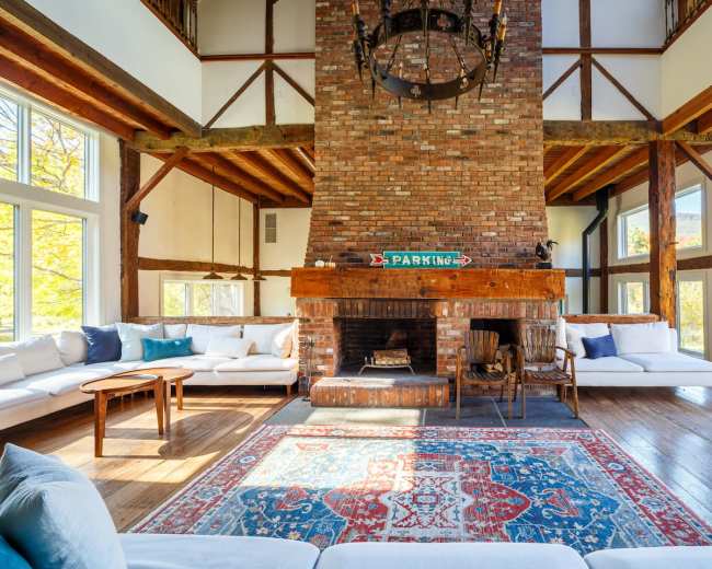 The image shows a spacious room with a large brick fireplace, wooden beams, and modern seating arranged around a decorative rug.