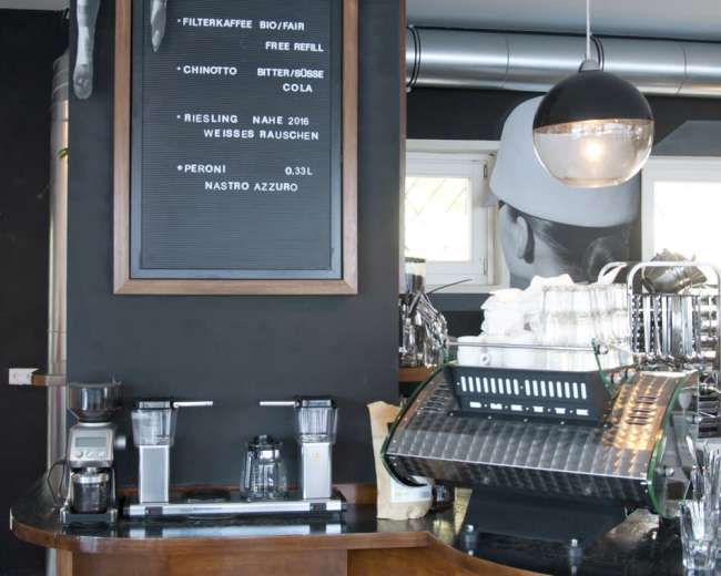 The image shows a modern coffee bar with a counter, coffee machines, and a chalkboard menu on a black wall.