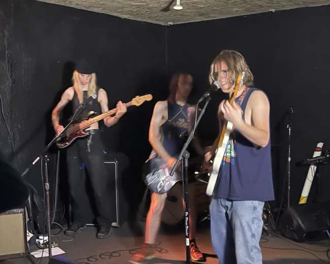 Three band members perform live on stage, with one playing a guitar and singing, while another plays bass, and the third is captured in motion.