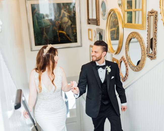 A bride and groom walk hand-in-hand down a staircase adorned with various decorative mirrors.