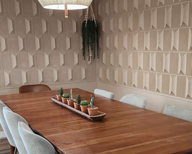 A wooden dining table with six upholstered chairs is set against a textured, patterned wall and features a pendant light above, along with small potted plants in the center.