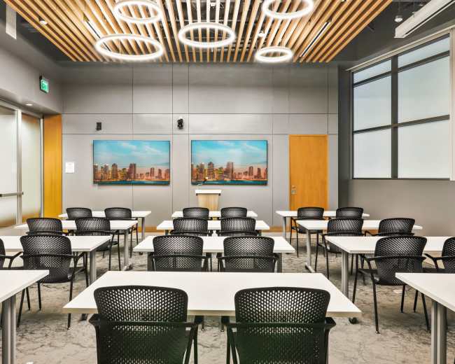 A modern classroom features rows of black chairs and white tables, with two framed cityscape artworks on the wall and a podium at the front.