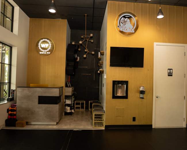 The image shows a modern fitness reception area with a front desk, wall-mounted television, and various gym equipment displayed in the background.