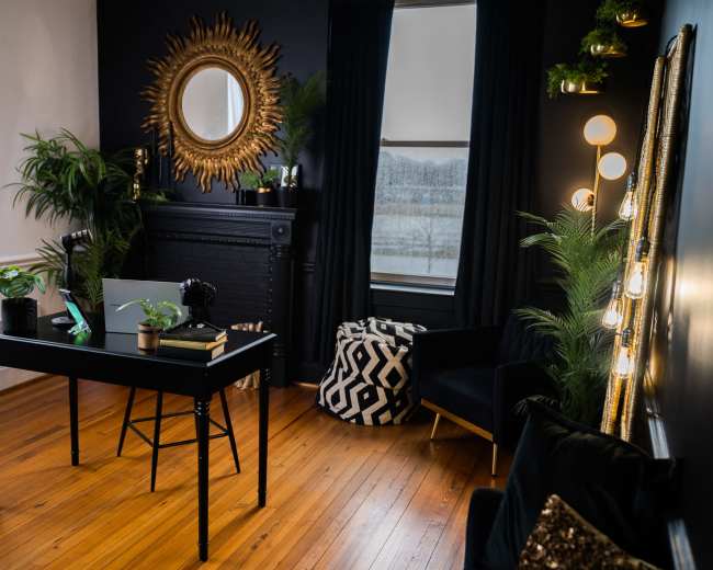 A modern office space features a black desk with a laptop, a ring light, decorative plants, and stylish furnishings against a dark wall.