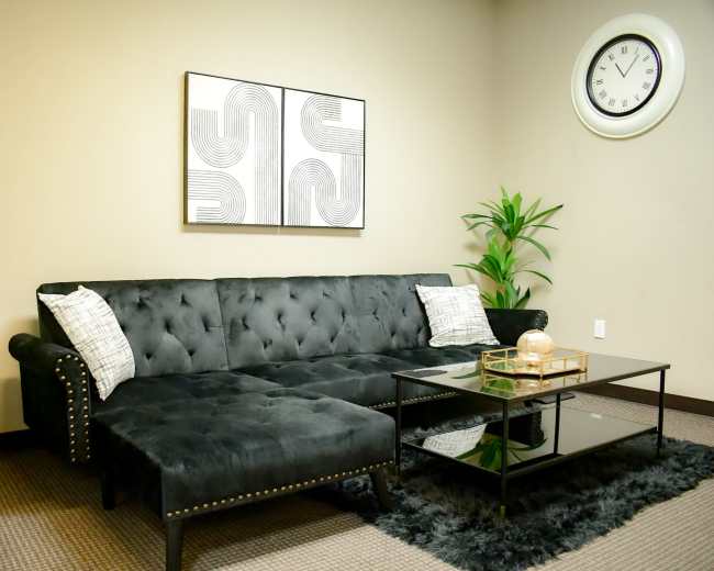 A modern living room featuring a black tufted sectional sofa, a glass coffee table, and decorative plants against a neutral wall.