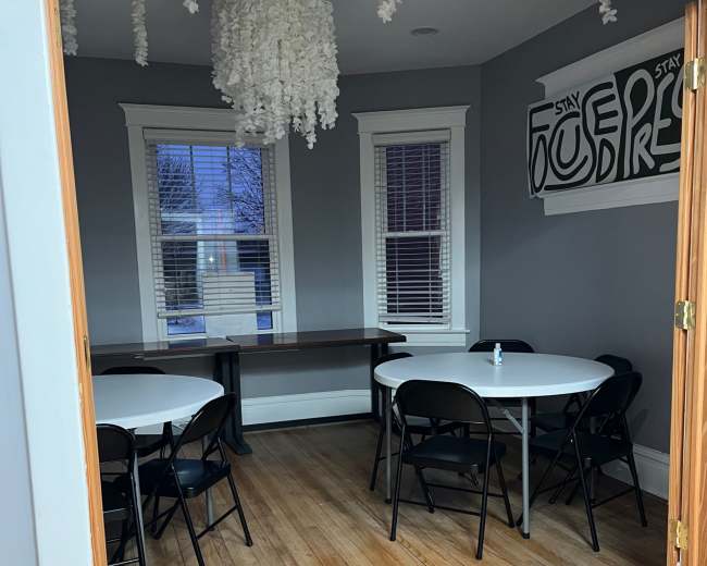 The image shows a room with gray walls featuring two round tables surrounded by black folding chairs, a wooden table along one wall, and a decorative light fixture hanging from the ceiling.