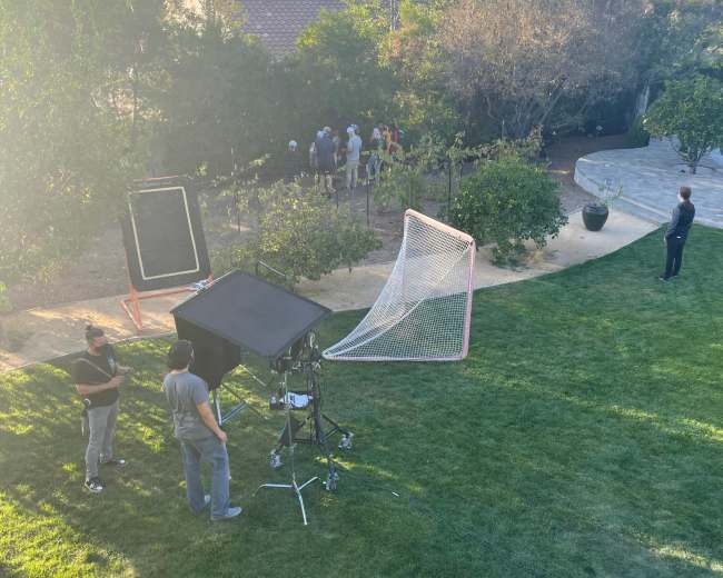 A film crew sets up equipment in a backyard while a group of people gathers near a bushy area.