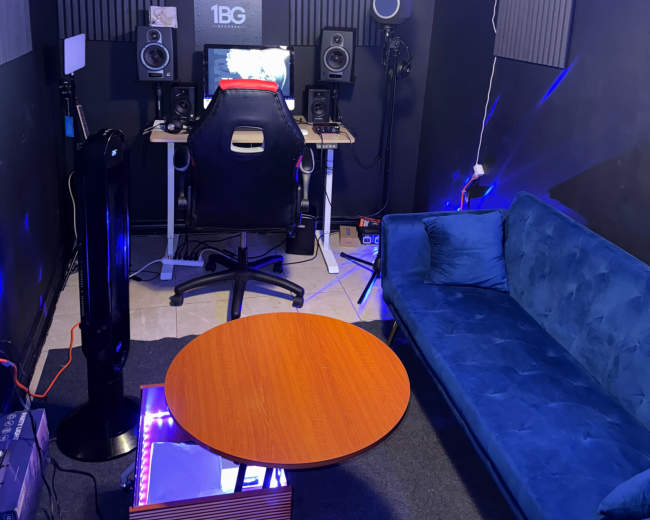 The image shows a small recording studio with a desk, computer, speakers, and a teal couch illuminated by LED lights.