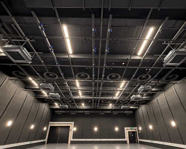 The image shows a spacious indoor room with a high ceiling adorned with ventilation units and overhead lighting.