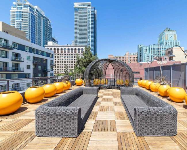 The image shows a rooftop lounge area with gray wicker furniture and large yellow planters, set against a backdrop of modern urban buildings under a clear blue sky.