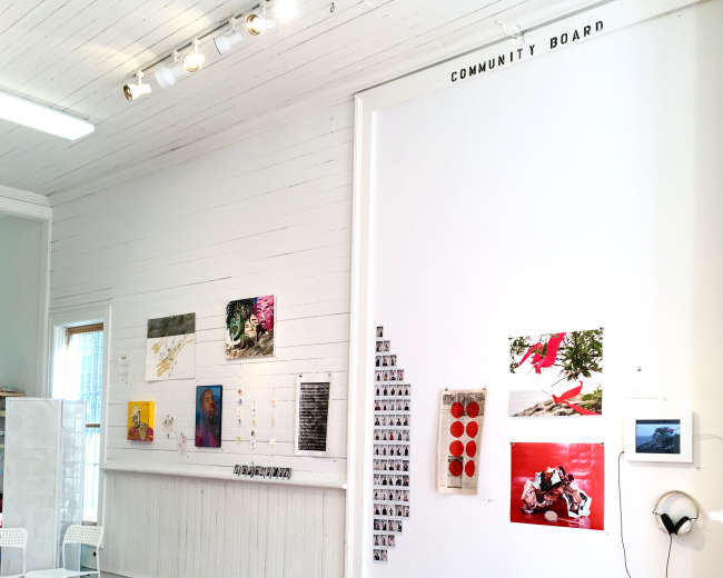 A gallery wall featuring a variety of artworks and a community board in a well-lit wooden room.