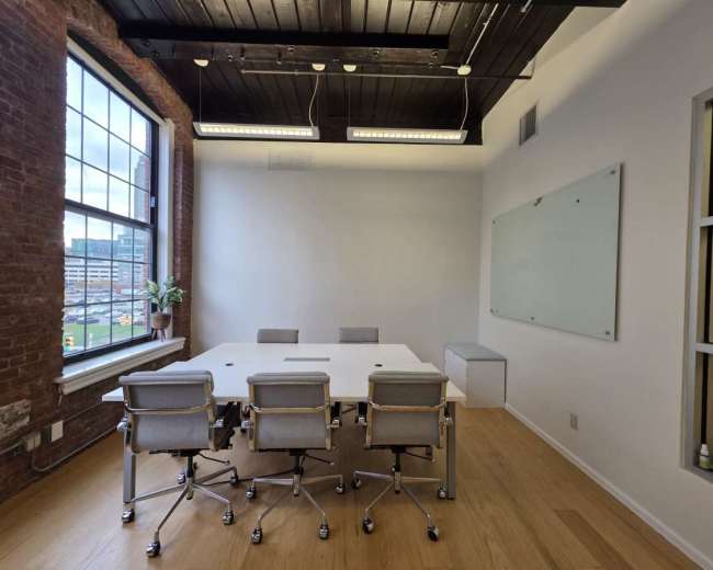 The image shows a modern conference room with a large white table surrounded by several ergonomic chairs, a whiteboard on the wall, and large windows revealing an outdoor view.