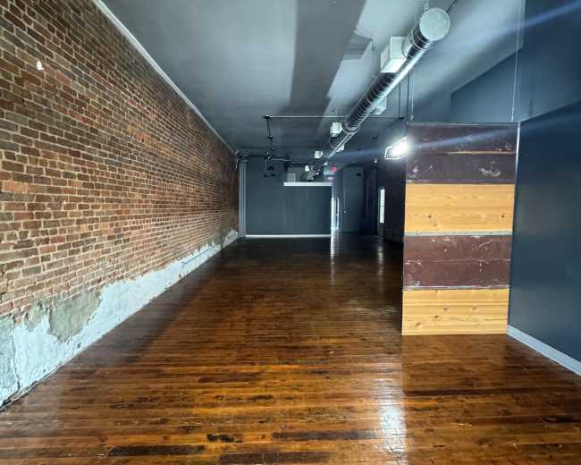 The image shows a spacious, empty room with hardwood floors, exposed brick walls, and a section featuring wooden paneling.