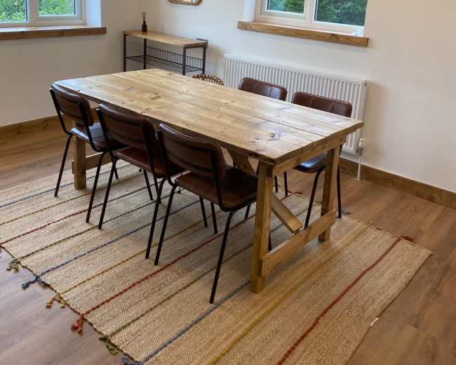 A rustic wooden dining table is surrounded by six chairs on a patterned rug, situated in a well-lit room with two windows.