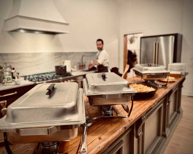 A buffet setup with metal chafing dishes is arranged on a wooden counter in a modern kitchen, where a man is preparing food in the background.