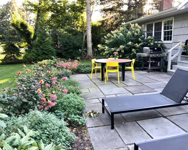 A patio area features a round table with yellow chairs, surrounded by a flower garden and a grill, adjacent to a house.